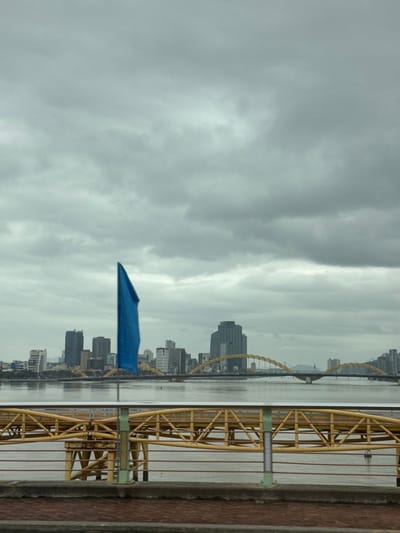 This image captures a wide-angle view of a cityscape and river under an overcast sky, located in Ngũ Hành Sơn Ward, Da Nang, Vietnam. In the immediate foreground, a metallic bridge railing featuring yellow structural elements and a silver handrail extends horizontally across the frame. Below the railing is a reddish-brown concrete surface. A single, vertically oriented blue flag, appearing folded or furled, stands on a pole to the left, rising from the railing into the mid-sky.
The midground is dominated by a wide, calm body of water, likely the Han River, reflecting the muted light from the sky. Across the water, in the background, a dense urban skyline is visible, characterized by numerous multi-story buildings, including modern high-rises and mid-rise structures. Spanning the river in the background is a prominent bridge with distinctive yellow, arched steel supports, identifiable as the Dragon Bridge. No individuals or active vehicles are visible in the scene. The sky is entirely overcast with uniform gray clouds.