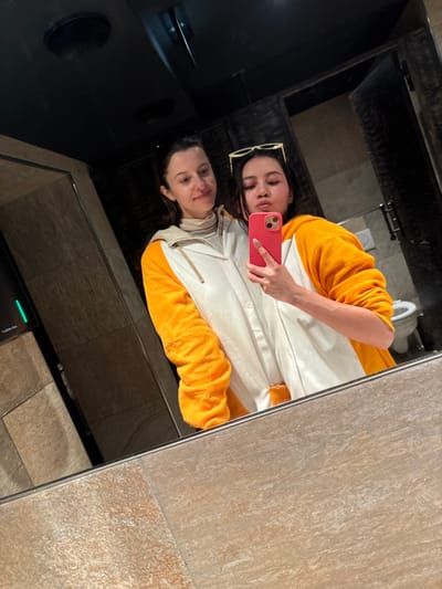 A mirror selfie captures a person in a white and yellow top, holding a pink phone. Sunglasses rest on their head. The reflection shows another image of the person, creating a double portrait effect. The bathroom background features tiles and a toilet. The dark mirror frame surrounds the reflective surface.