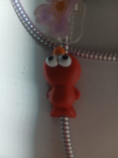 A blurry, close-up photograph shows a red, cartoon-like figure with large, white eyes and orange accents on its head, dangling from a metallic, coiled cord. A blurry, light purple flower with an orange center is visible behind the figure and cord. The background is a light, reflective surface, possibly a table or countertop, with some dark specks that might be dust or debris. The image lacks sufficient detail to determine the location in Navaluenga, Spain, or any specific actions or interactions.