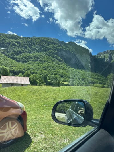 A red car is parked on a grassy area with a building and a mountain in the background. The image appears to be taken from inside the car, looking out the passenger side window. The car's side mirror reflects a person holding a phone and wearing a ring. The building is stone with a red tiled roof, and it is surrounded by trees. The mountain is covered in lush green trees and has steep rocky cliffs. The sky is blue with white clouds. The location is Vusanje, Montenegro.