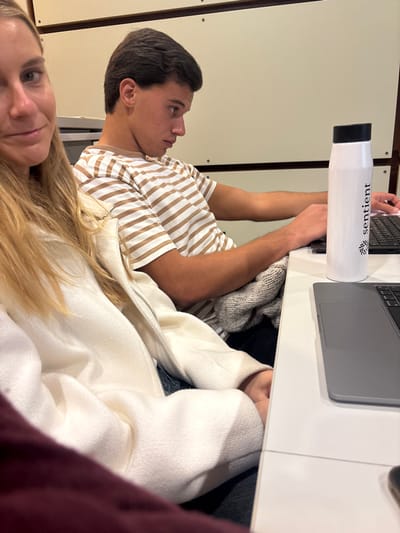 This image captures an indoor scene, likely a workspace or study area, in Porto Salvo, Portugal. In the foreground, partially visible on the left, is a female with long blonde hair and light skin, wearing a light-colored, possibly white, zipped jacket. Her right eye, nose, and part of her mouth are visible, and she appears to be looking towards the camera with a slight smile. To her right, a young adult male with dark hair and a striped t-shirt (alternating light brown/beige and white stripes) is seated. He is viewed in profile, facing right, and is focused downwards, actively typing on a dark laptop with both hands. A white cylindrical water bottle with a black lid, bearing the word "sentient" and a logo, stands upright on the white table between his hands and the edge of another, partially visible dark grey laptop. A light grey knit garment is draped over his lap. The background consists of light-colored paneled walls with dark horizontal lines and visible circular fasteners.
