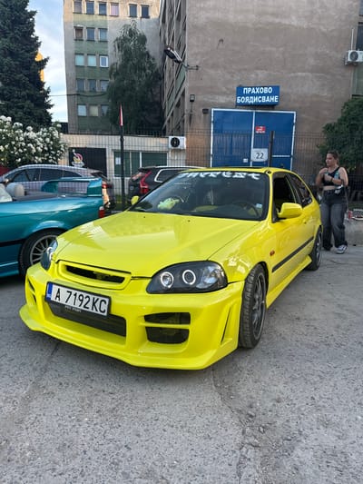 A bright yellow modified hatchback, a Honda Civic, is parked on asphalt next to a teal convertible. In the background, a woman in a black crop top and jeans stands near a blue garage door with Bulgarian signage indicating "ПРАХОВО БОЯДИСВАНЕ" (Powder Coating). The building behind the garage appears to be an industrial structure with multiple windows. A small white number "22" is visible on the garage door. The scene is set in Busmantsi, Bulgaria, likely in an industrial or commercial area.