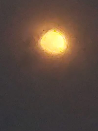 A photograph of a luminous yellow circle against a dark, textured background. The circle appears to be a light source, possibly a lamp or a celestial body, with a halo effect. The background has a grainy texture, suggesting it could be a wall or a sky. The image is abstract and lacks specific identifiable objects or subjects. There is no discernible location in Kyzylorda, Kazakhstan.