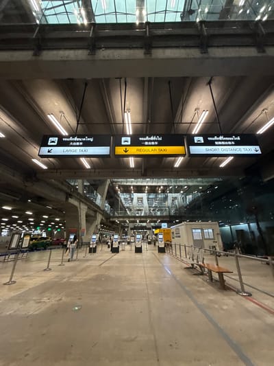 This media file captures a scene inside an airport or transportation hub in Nong Prue Subdistrict, Thailand. The image is oriented sideways, presenting a perspective looking down a wide, well-lit corridor. The primary subjects are signage indicating taxi services.
There are three prominent signs, stacked vertically, all featuring text in Thai and English, along with some Chinese characters. The top sign reads "SHORT DISTANCE TAXI," the middle sign indicates "REGULAR TAXI" with a downward-pointing arrow, and the bottom sign says "LARGE TAXI" with a leftward-pointing arrow. These signs are mounted on a dark structure that runs parallel to the main walkway.
The setting appears to be an indoor area with a high ceiling, illuminated by numerous strip lights. To the left, a large glass facade allows some natural light to enter and reflects the interior. To the right, a concourse area is visible with various kiosks or service counters, some displaying information on screens. A few people are present in the distance, appearing to be passengers or staff.
The overall impression is that of a functional, modern transportation hub. The lighting suggests it could be evening or a dimly lit indoor space. There are no explicit indications of weather, but the interior setting implies protection from the elements. The scene is orderly and designed for passenger flow, with clear directional signage to guide travelers to different taxi services. The visible text provides clear instructions for accessing taxi transportation.
