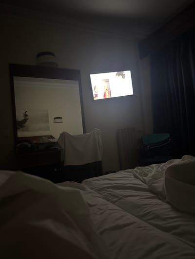 The image depicts a dimly lit hotel room with a large unmade bed in the foreground. To the left, a large mirror reflects part of the room, showing a desk with items on it and a piece of artwork on the wall. A white chair draped with fabric is visible in the middle ground, positioned near the mirror. A television screen is on the wall to the right, displaying a bright image. A dark blue curtain is drawn on the far right side of the room, and a white radiator or heating unit is situated below the television. A dark-colored suitcase and a teal bag are placed on a chair to the right of the radiator. The overall impression is of a lived-in space, likely a hotel room in Fuengirola, Spain, given the context provided.
