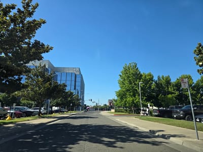 This is a daytime, eye-level, outdoor, wide shot captured from a street in Sacramento, California. The scene depicts a street lined with trees, cars parked on the side, and a modern office building with blue glass windows. The sky is clear and bright blue. The street is paved and has visible lane markings. Shadows of trees are cast across the road. On the left side of the road, a large office building with a white and blue facade is visible. Trees are planted along the sidewalk in front of the building, and several cars are parked. On the right side of the road, more trees and parked vehicles are present. A "NO PARKING ANY TIME" sign is mounted on a pole. Traffic lights are visible in the distance at an intersection. A person holding a pink umbrella is walking across the street in the distance. The overall impression is a suburban or business park setting.