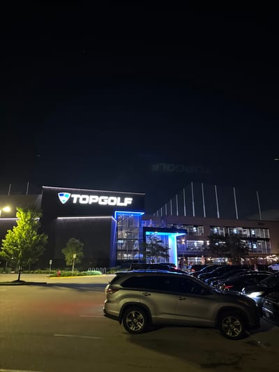 An exterior nighttime shot captures the Topgolf entertainment venue in Schaumburg, Illinois, USA. The building, marked with a prominent "TOPGOLF" sign illuminated in white and blue, is silhouetted against a dark sky. In the foreground, a silver SUV is parked in a lot, with other vehicles visible behind it. The scene is lit by ambient light from the venue and streetlights, casting shadows and highlighting the architectural features of the building and the surrounding trees. The topgolf nets are visible behind the main structure, indicative of the venue's purpose.