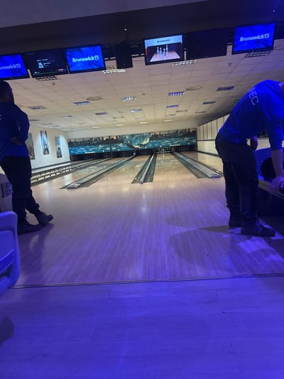 The image shows the interior of a bowling alley. Two individuals are visible, one preparing to bowl and the other standing towards the end of the alley. The bowling alley features multiple lanes, each with a Brunswick scoring screen overhead. The walls are adorned with images or designs and the lighting casts a blue hue. No information to confirm the location being in Gioia Tauro, Italy.