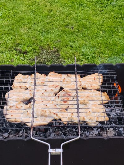 The image shows chicken pieces cooking on a grill. The chicken is arranged in a grill basket, with several pieces laid out horizontally. It appears to be seasoned with herbs. The grill is over hot coals, with some flames visible on the right side and a wisp of smoke rising. The grill itself is black and sits within a larger black charcoal grill. The background is out of focus but appears to be a grassy lawn with some patches of dirt or dried grass. The scene suggests an outdoor setting, possibly a backyard or picnic area in Russia, given the location context provided. The lighting indicates it is daytime, likely not direct midday sun given the slight shadows. There is no visible text in the image. The overall impression is of a meal being prepared outdoors.