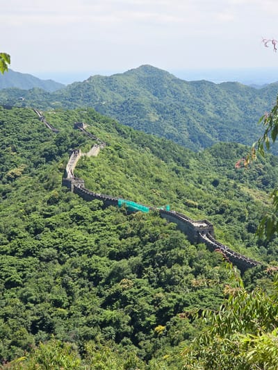 The image shows a section of the Great Wall of China winding along a lush, green, mountainous terrain in Huairou District, China. The wall, constructed from stone and brick, is visible with watchtowers and battlements. Numerous tourists are walking on the wall, some grouped together and others spread out. The surrounding landscape is densely covered with trees, with rolling hills and mountains extending into the distance. The sky is overcast with a hazy, pale blue. In the foreground, leafy branches partially obscure the view, suggesting the perspective is from a higher vantage point. A section of the wall in the middle ground appears to be under some form of repair or preservation, indicated by green netting.