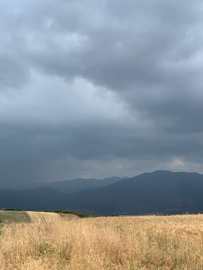 This is a vertical, eye-level, medium shot of a landscape in Γαλαρινός, Greece. The scene depicts a transition from a storm-laden sky on the left to a sun-drenched field of dry grass on the right. The sky is filled with dark, heavy clouds, suggesting an impending or ongoing storm. In the distance, layered mountain silhouettes are visible, gradually becoming darker and more obscured by the stormy weather as they recede. A clear line separates the dramatic sky from the mountains and the bright, golden field of dry grass that occupies the right side of the frame. The field appears to be on a gentle slope, with a path or tire tracks visible across it. The overall impression is one of atmospheric contrast, with the brewing storm on one side and the warm, dry landscape on the other. The time of day is not explicitly clear, but the lighting suggests it could be late afternoon, with the sun trying to break through or having just passed over the mountains.