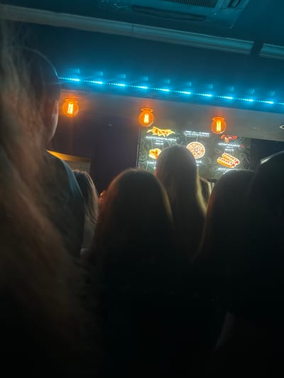 The image appears to be taken inside a dimly lit establishment, likely a cafe or fast-food restaurant, in Kirov, Russia. The scene is characterized by the presence of several individuals, their faces mostly obscured, suggesting they are patrons. In the background, a digital menu board is visible, displaying images of food items like french fries, pizza, and hot dogs, along with prices in Russian rubles. The menu board is illuminated by warm-toned Edison-style light bulbs hanging from above, and further accentuated by a strip of bright blue LED lights running horizontally across the upper portion of the board. The overall ambiance suggests a casual, possibly nighttime setting.