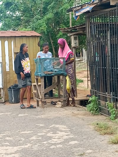 Three women are gathered around a small wooden table with a wire mesh cage on top. One woman, wearing a pink head covering and a patterned dress, stands at the table. Another woman in a striped shirt is also at the table. The third woman, wearing a black sweatshirt with a cartoon graphic and jean shorts, stands to the left of the table. A black trash can is visible behind her. The setting is outdoors, near a road with a small wooden building to the left and a structure with metal bars to the right. Green trees are in the background. This is in Oshogbo, Nigeria.