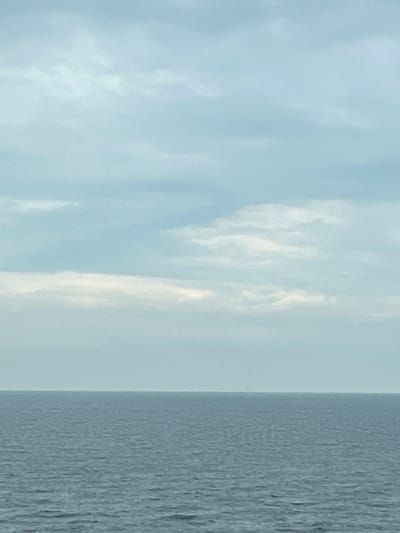 A vast, calm, gray-blue ocean stretches to the horizon under a pale blue sky filled with wispy, white clouds. The horizon line is indistinct, suggesting a hazy atmosphere or a distant, unidentifiable object on the horizon. The water is mostly smooth with subtle ripples, indicating minimal wave activity. The overall impression is one of a serene and expansive seascape. The location is Unknown, Malaysia.