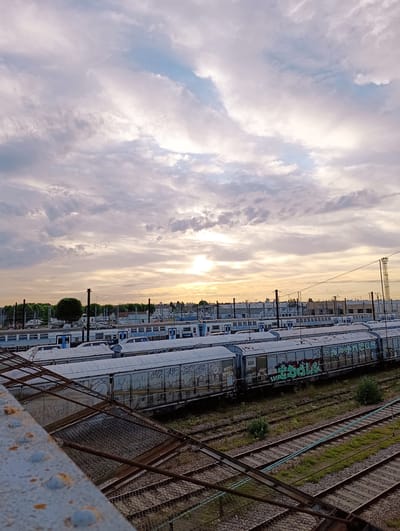 The image depicts a railway yard in Villeneuve-Saint-Georges, France, under a cloudy sky at sunset. Several trains are parked on multiple tracks. The trains appear to be passenger cars and cargo wagons, some of which are covered in graffiti. In the background, there are buildings, trees, and utility poles. The foreground shows a metallic structure with a wire mesh, likely part of a walkway or maintenance area, with a rusted metal railing. The overall scene suggests an industrial area during the evening, with the soft light of the setting sun casting a warm glow on the clouds and the trains. There are no people visible in the image.