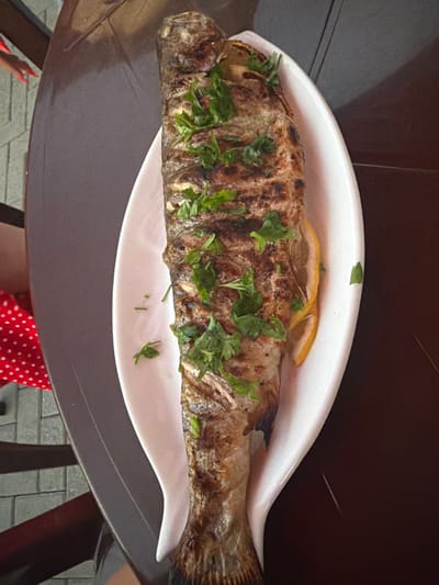 A whole, grilled fish, garnished with parsley and lemon slices, is presented on a white oval platter. The platter rests on a dark, circular table. To the left of the table, a person wearing a red and white polka dot dress is visible, with a hand featuring red nail polish reaching towards the table's edge. The background shows a tiled surface and the edge of what appears to be a restaurant or outdoor seating area. The scene is located in Amzara, Abkhazia.