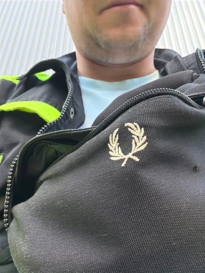 A close-up, low-angle shot shows the chest area of an unidentified person wearing a black jacket with a light blue t-shirt underneath. The jacket has a prominent neon yellow accent strip and a zippered opening. A laurel wreath logo, embroidered in a light beige or gold thread, is visible on the left side of the jacket. The background consists of a ribbed, light-colored wall, likely an exterior surface. The image appears to be a selfie, focused on the clothing and the person's chin and neck. There is no visible indication of the location being Syktyvkar, Russia.