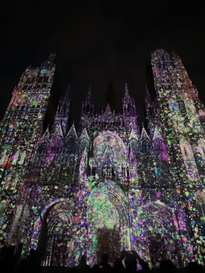 The image displays the facade of Rouen Cathedral (Cathédrale Notre-Dame de Rouen) in Rouen, France, at night. The Gothic architectural structure is illuminated by a dynamic, multi-colored light projection. The projection features numerous small, irregular shapes resembling paint splatters or confetti, primarily in shades of purple, green, blue, pink, and yellow, distributed across the entire surface of the cathedral. Two large, ornate towers rise on the left and right sides of the main facade, with intricate spires and arched elements visible in the central section. At the base of the image, several human silhouettes are discernible, indicating an audience viewing the projection. Some individuals appear to be holding up mobile devices, presumably to photograph or record the event. The sky above the cathedral is completely dark and featureless.