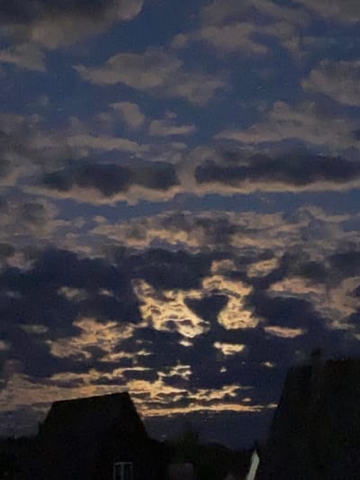 The image presents a low-light outdoor scene in Zheleznodorozhnyi, Russia, characterized by a prominent cloudscape and the dark silhouettes of architectural structures. The sky occupies the majority of the frame, displaying a complex pattern of fractured and stratified clouds. The upper portions and bodies of the clouds appear dark blue to grey, while extensive areas of their undersides and edges are brightly illuminated with hues of yellow, orange, and light brown, suggesting a light source positioned below or behind the cloud layer, likely during twilight or dawn.
The lower third of the image features the dark, undifferentiated silhouettes of at least two building rooftops. On the left, a distinct gabled roof is visible, extending diagonally upwards towards the center, with a faint rectangular shape, possibly a window, discernible beneath its peak. On the right, a partial silhouette of another sloped roof structure is present, its form less defined, with a vertical light-colored line visible on its leading edge. No individuals, animals, or specific actions are discernible within the frame. The environment indicates a residential or suburban setting under crepuscular or nocturnal conditions.