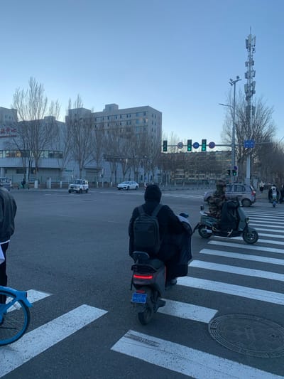 A person is riding a moped across a crosswalk in an urban area. The rider is wearing a black jacket and a backpack. Another moped is stopped nearby. Traffic lights are visible in the background and show green. There is a modern building and trees without leaves. A tall structure with numerous cameras is also visible, with a clear blue sky in the background.