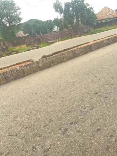 The image shows a road in Kona Garu, Nigeria. The road is paved and has a curb made of concrete blocks. On the left side of the road, there are some houses with thatched roofs and corrugated iron roofs. There are also some trees and vegetation. On the right side of the road, there is a house with a corrugated iron roof and a wall. The sky is overcast. The image appears to have been taken from a low angle, looking across the road.