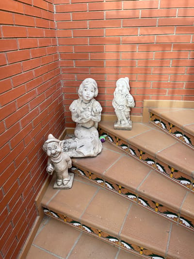 The image depicts a corner of a courtyard or outdoor staircase area, with a red brick wall on the left and a set of tiled stairs on the right. Three garden gnome-like statues are placed on the steps and at the base of the stairs. One statue, resembling Snow White, is seated on the first step, holding a small animal. To her left, a smaller gnome statue with a hat and a wide smile is standing at the foot of the stairs. Further up the stairs, another gnome statue in a standing position is visible. The stairs are made of terracotta-colored tiles with a decorative border featuring a pattern of flowers and leaves. A small, colorful ball is lying on the step near the seated statue. The scene is lit by natural light, casting shadows on the wall and stairs. The location is identified as Santa Coloma de Gramenet, Spain.