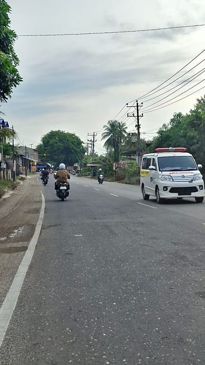 An asphalt road in Langa, Indonesia, features several motorcycles and an ambulance. The road has a white painted line on the left side, marking the edge of the paved surface. In the distance, several buildings line the left side of the road, some with simple structures and corrugated roofs. Tall trees and utility poles with power lines are visible throughout the scene, suggesting a semi-urban or suburban environment. The sky is overcast with gray clouds.

On the road, a person wearing a white helmet and a patterned shirt is riding a scooter, traveling away from the viewer. To the left and further ahead, two other motorcycles are visible. On the right side of the road, a white ambulance with red lights on its roof is driving in the same direction as the motorcycles. The ambulance has a yellow stripe and text that appears to read "AMBULANS" in reverse. The road surface is textured and shows signs of wear and tear. Green foliage and trees are present on both sides of the road.