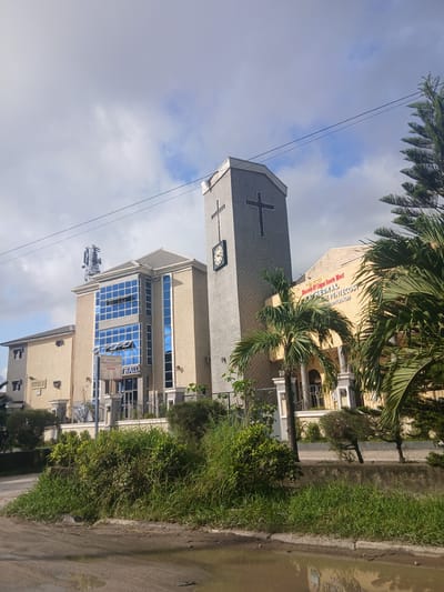 A wide shot captures the exterior of a church in Satellite Town, Nigeria, under a partly cloudy sky. The foreground is dominated by a muddy, waterlogged dirt road reflecting the surrounding environment. Lush green vegetation, including tall grasses and bushes, lines the edge of the road and the church property.
The main subject is a multi-story church building. The central structure is a tall, gray tower with a clock face and a prominent black cross. Adjacent to the tower is a lighter-colored building with multiple floors and large blue-tinted windows. A sign above the entrance reads "CATHEDRAL CHURCH OF THE PENTECOST" and "HALL." Another building, also lighter in color, is visible to the left. The church appears to be an active place of worship, indicated by its signage and architectural style. The surrounding environment suggests a tropical or sub-tropical climate, given the vegetation. No people are visible in the frame.