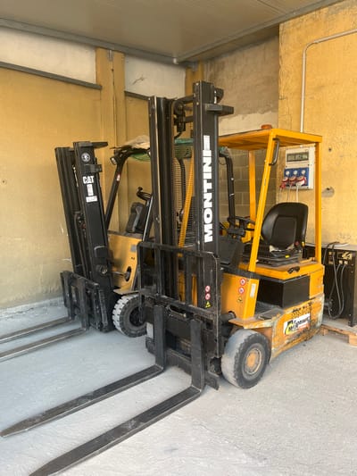 Two forklift trucks are parked next to each other in an outdoor, covered area with a concrete floor and yellow and grey walls. The forklift on the right is yellow and black and has the brand "MONTINI" visible on its mast. It has a black seat and a control panel with electrical outlets attached to its frame. The forklift on the left is black and has the "CAT" logo visible on its mast. Both forklifts have their forks extended forward and resting on the concrete floor. To the right of the forklifts, there is a black electrical box with wires connected. The scene appears to be a storage or maintenance area, possibly in an industrial or commercial setting within Rozzano, Italy. There are no people present in the image.