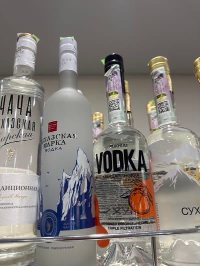 This is a close-up, vertical shot of several bottles of alcohol displayed on a metal shelf in what appears to be a retail setting. The bottles are predominantly glass and contain clear or light-colored liquids, likely vodkas or other spirits. Some bottles have decorative labels with images of mountains, while others feature fruit motifs, such as an orange on one label.

The visible text on the labels is primarily in Cyrillic and English, indicating brands, ingredients, and production details. Some of the text mentions "VODKA," "SUKHUM," "PREMIUM QUALITY," and "TRIPLE FILTRATION." There are also phrases in Cyrillic that can be translated to "Abkhazian," "MANDARIN," and "PHEASANT." One label specifies the location as "PROIZVEDENO V ABKHAZII," meaning "Produced in Abkhazia." Another label reads "SUKHUM," which is a city in Abkhazia.

The lighting suggests an indoor environment, likely a store, with artificial lighting. The overall impression is a display of alcoholic beverages, with a focus on their branding and origin from Abkhazia. There are no people or specific events depicted; the scene is static and focuses solely on the products.