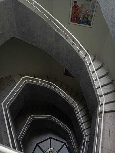 This is a high-angle, downward view of a spiral staircase in a building. The staircase has white railings and appears to be made of marble and dark concrete. There are at least four levels visible, descending into darkness. On one of the upper walls, there is a framed painting depicting two men, one playing a horn and the other an accordion. The floor of the staircase on the upper levels has square tiles. The location is an interior space, likely a public building or residence, in Constance, Germany. No individuals are present or actively interacting within the frame of the image.