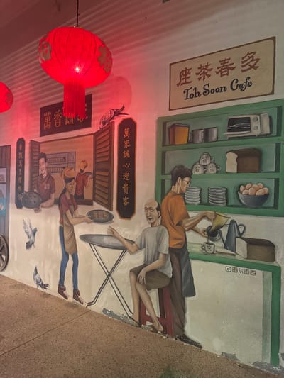 This image depicts a mural on a light-colored wall, illuminated by the red glow from two real-world, hanging red Chinese lanterns with tassels. The mural is located in George Town, Malaysia, specifically featuring a scene from "Toh Soon Cafe," a name prominently displayed on a sign at the top right in both English and Chinese characters.
The mural illustrates a traditional cafe scene with multiple figures and objects. On the left, a smiling male cafe worker in a red shirt holds a tray of baked items, visible through an open, brown-shuttered window. Another worker is partially visible behind him. Below this, two grey pigeons are depicted on the mural, one in flight, one walking on the ground level.
In the center, a female worker wearing a red shirt, blue jeans, and an apron extends a tray holding three small, round baked items towards an elderly, bald male customer. This customer, dressed in a light grey t-shirt and brown shorts, is seated on a red stool at a small, round, grey table with an X-shaped base. He gestures with his left hand towards the approaching tray.
On the right side of the mural, a male worker in an orange t-shirt and dark apron stands behind a green counter, actively pouring liquid from a traditional coffee sock filter into a white cup. Above this counter, green shelves display various cafe items: two yellow containers, two grey mugs, a stack of white patterned cups, a white toaster oven, a loaf of bread, and a bowl of brown eggs. A stack of white plates is also visible on the counter. A pigeon is perched on a brown shutter above the central interaction.
Vertical Chinese calligraphy signs are painted on the wall on the left and center. The ground in the foreground is a paved, light brown pathway.