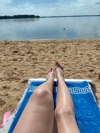The image shows a first-person perspective of a person relaxing on a beach lounger on a sandy beach. Their legs, with red-painted toenails, are extended and resting on a blue towel with a white geometric pattern. The sand is a light brown and appears to be lightly textured with footprints.
In the background, a calm body of water, likely Lake Myadel, stretches out to the horizon. A thin line of trees marks the distant shore. The sky is blue with scattered white clouds. A small bird is visible in flight over the water. A pink flip-flop is partially visible on the left side of the lounger. The scene suggests a sunny day at a lakeside beach in Myadzyel, Belarus.