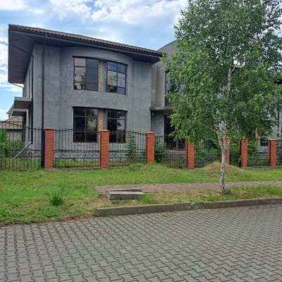 The image depicts a residential property in Tairove, Ukraine. The house is under construction, with unfinished grey stucco walls and exposed brickwork around some of the windows. The roof is tiled with brown shingles, and there are dark-framed windows visible. A sign with Ukrainian text, including "ПРОДАМ" (for sale), is displayed in one of the upper windows, along with a QR code. A black wrought-iron fence with decorative elements, supported by brick pillars, separates the property from the street. In front of the fence, a patch of grass is visible, and a small birch tree stands to the right of the house. The foreground shows a paved area made of interlocking grey concrete pavers, with a concrete curb separating it from the grassy area. The sky is cloudy.