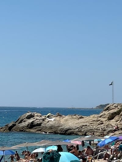This is a vertical, medium-shot photograph taken on a sunny day at Platja Gran, a beach in Platja d'Aro, Castell d'Aro, Spain. The image captures a section of the coastline with a rocky outcrop in the middle distance and a crowded beach in the foreground. The sky is a clear, pale blue, taking up the top half of the frame. The sea is a vibrant turquoise and appears calm, with gentle waves lapping at the shore.
In the middle ground, a large, weathered rock formation dominates the scene, jutting out into the sea. Two light-colored towels or cloths are draped over the rock. Further in the distance, a thin, dark flag on a tall flagpole stands on a rocky promontory to the right. The sea extends to the horizon, where a faint line of land or structures is visible.
The foreground of the image is filled with a section of a beach, teeming with activity. Numerous people are spread out on the sand and in the shallow water, many of them relaxing under colorful beach umbrellas. Beach chairs and towels are visible, suggesting a busy summer day. The people are mostly seen from the torso up, with some lying down and others standing. The umbrellas are a mix of colors, including blue, pink, and white, creating a festive atmosphere. The water at the edge of the beach is shallow and clear, allowing a glimpse of the sandy bottom. The overall impression is one of a vibrant and popular tourist destination.