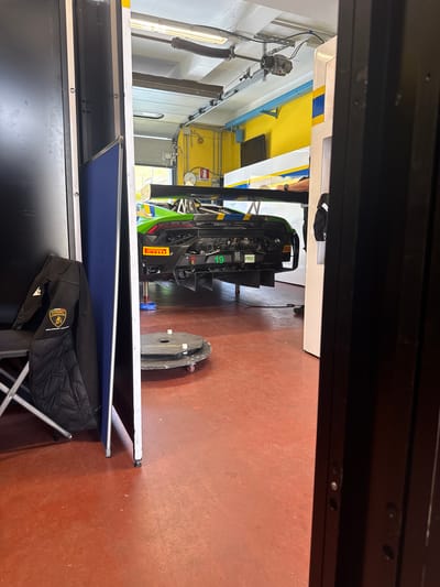 The image captures a scene inside a garage or pit area, likely in Campagnano di Roma, Italy, given the Lamborghini racing equipment present. A green and black Lamborghini race car is visible towards the back of the space, possibly undergoing maintenance or adjustments. There's a large circular wheel or component of some sort near the car. The floor is a reddish-brown color. In the foreground, a table is visible with a blue fabric surface, and a black Lamborghini-branded bag is resting on a folding chair next to it. The photo is taken from the perspective of someone looking into the space through a doorway or opening. Overhead structures include lighting and mechanical systems, painted in white, yellow, and blue.