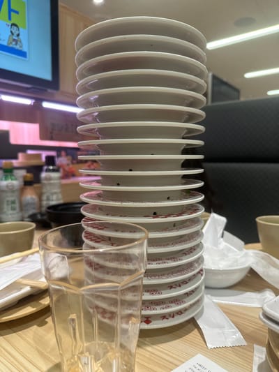 The image depicts a dining table within a Sushiro conveyor belt sushi restaurant in Nakano, Japan. The central focus is a tall, vertical stack of approximately 20-22 used white sushi plates, each featuring a red geometric pattern and the Japanese text "スシロー" (Sushiro) on its underside. Some plates have small, circular holes, likely for an automated plate counting system.

In the immediate foreground, a clear, faceted drinking glass stands to the left of the plate stack. To the left of the glass, a pair of wooden chopsticks rests on a small, light-colored wooden tray, with white paper wrappers. A light brown ceramic cup is also visible to the left.

To the right of the stacked plates, a small white bowl is partially obscured by crumpled white paper or a napkin, and another light brown ceramic cup is present. White paper packets, possibly containing wet wipes or condiments, are scattered on the light wooden table surface.

The background shows elements of the restaurant environment. On the left, a television screen displays an animated character on a blue background. Beyond the table, a raised counter area features several bottles of condiments, including what appears to be soy sauce. Horizontal fluorescent lighting illuminates the space. Dark, possibly upholstered, booth seating is visible on the right side of the frame. Blurred figures of individuals can be discerned in the deeper background, indicating other patrons or staff.