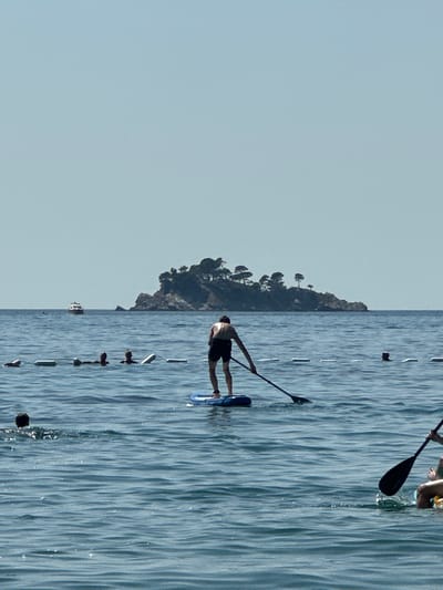 The image shows a body of water with a few people engaged in water activities. In the foreground, a person is paddleboarding and rowing with a paddle. To the left, other individuals are in the water, some with their heads visible. A floating barrier with buoys is present in the water, marking an area. In the background, a small island covered with trees and rocks is visible. A boat is also seen in the distance. The sky is clear and blue. The scene appears to be a beach or coastal area, likely in the vicinity of Đurovići, Montenegro, given the rocky island formation and clear water.