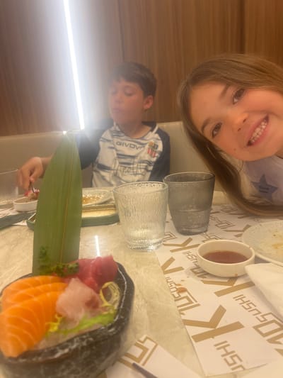 A child is seated at a table in a restaurant, possibly in Torreforta, Spain. Two children are visible in the image. The table has sushi dishes, beverages in glasses, and a small bowl, possibly with soy sauce. The table covering is patterned with the word "SUSHI". The restaurant appears to have light-colored wood paneling.