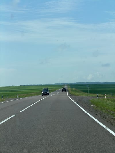 The image depicts a straight, asphalt road stretching into the distance under a light blue sky with scattered clouds. The road has white lane markings. To the left and right of the road are expansive, vibrant green fields, suggesting a rural or agricultural setting. There are several cars visible on the road; a black SUV is in the foreground on the left lane, and another black car is further ahead in the right lane. Roadside markers, some with red and white reflective elements, line the edges of the road. A blue signpost with white lettering is visible on the right side of the road in the distance. The horizon is marked by a line of trees and rolling hills. The overall scene conveys a sense of travel and open landscape. There are no visible buildings or specific landmarks that would pinpoint the location within the city of Krasnaya, Russia, based solely on the visual information provided.