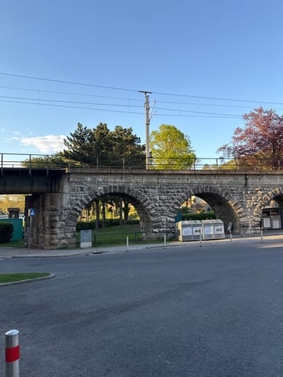 This image depicts a sturdy, multi-arched stone railway viaduct, characteristic of architecture found in regions like Hainburg an der Donau, Austria. The viaduct, constructed from rough-hewn stone blocks, spans over a paved road and grassy areas.
The scene is bathed in warm, late afternoon or early evening sunlight, creating distinct shadows and illuminating the textures of the stone. The sky is clear and bright blue, with a few faint clouds visible on the left.
In the foreground and to the right, an asphalt road runs alongside the viaduct. Beneath one of the arches, several light-colored recycling bins are neatly arranged. Clearly legible German text on these bins includes "VERPACKUNGEN" (packaging), "PAPIER" (paper), and "GLAS" (glass), accompanied by corresponding pictograms. A blue pedestrian crossing sign with a white 'A' symbol is visible near the left arch, indicating pedestrian access. Red and silver bollards mark the road's edge further down.
A single person, dressed in blue, can be faintly seen in the grassy area underneath an arch, near the recycling bins, suggesting ordinary daily activity. Overhead, a tall utility pole with multiple power lines stretches across the left portion of the sky. Various trees with green and reddish-brown foliage are scattered around the viaduct, contributing to the natural elements of the urban landscape. The overall impression is one of a peaceful, well-maintained public space, integrating historical infrastructure with modern utilities.