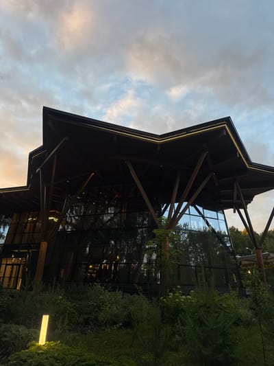 The image depicts a modern building with a distinctive, angular roof supported by numerous angled wooden beams. The building features large glass panels that reflect the surrounding trees and the twilight sky, which is filled with scattered clouds tinged with hues of orange and blue. In the foreground, lush green foliage and a tall, rectangular outdoor light fixture create a naturalistic setting. The architecture suggests a contemporary design integrated with its environment. The building is likely located in or near the city of Sosny, Russia, given the context.