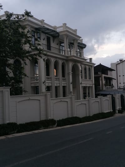 The image captures a grand, cream-colored villa with classical architectural elements, located in Xuan Truong, Da Lat, Vietnam. The villa features multiple stories, arched windows, balconies with ornate railings, and a decorative balustrade along the rooftop. Flanking the villa are lush green trees and a meticulously crafted white fence. The scene is set against a backdrop of a cloudy sky, suggesting it might be late afternoon or early evening. The foreground shows a paved road, indicating a street-side view of the property. Adjacent to the main villa, other buildings of varying architectural styles are visible, one with a dark, sloping roof. The overall impression is one of opulent residential architecture in a potentially affluent neighborhood.