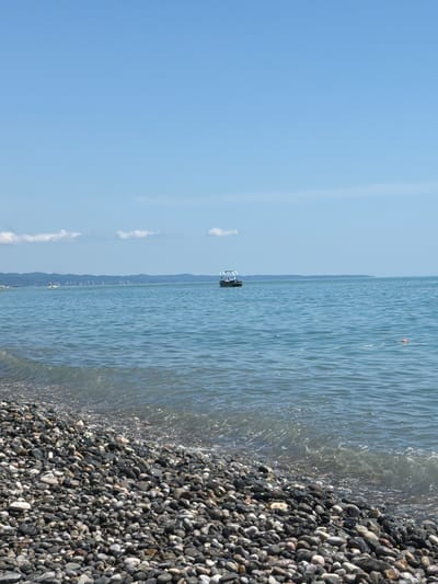 A wide shot of a pebbly beach in Tsandrypsh, Abkhazia, shows a clear blue sky above a tranquil, light-blue sea. A small, dark boat with a canopy is visible in the distance on the water, to the right of a distant, hazy shoreline with a range of mountains. In the foreground, the beach is covered in smooth, gray, and dark-colored pebbles. Small waves gently lap at the shore, creating a foamy edge where the water meets the stones. There are no people visible in the frame.