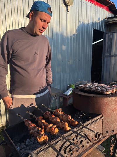 The image depicts a man wearing a blue baseball cap and a grey sweatshirt, standing in front of a barbeque grill. The grill is filled with skewers of meat, which are being cooked over hot coals. In the background, there is a corrugated metal structure, likely a shed or garage, with an open doorway. A table and some bags are visible to the left of the grill. The scene appears to be outdoors, with sunlight casting shadows on the ground and the metal siding. The location is identified as the village of Dunilovo, Russia.
