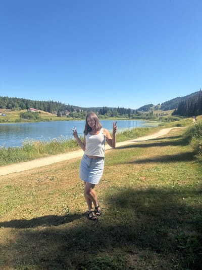 A woman stands on a grassy area next to a path, facing the camera and making a peace sign with both hands. She is wearing a white tank top and a light blue denim skirt, with black sandals. Behind her is a lake with calm water, bordered by green hills and trees. Several buildings are visible on the slopes of the hills. The sky is clear and blue. The scene appears to be a sunny day in a rural or park-like setting. This location is in Lamoura, France.