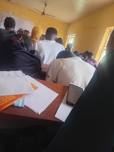 The image depicts an indoor scene within a classroom setting, likely in Ogbogbo, Nigeria, based on contextual information not visually present in the image. The room is characterized by its yellow-painted walls and ceiling, with fluorescent lighting fixtures and a ceiling fan visible. Sunlight streams in through two windows on the right side of the frame, illuminating the space.
Several individuals, primarily young adults or teenagers, are seated at wooden desks, facing towards the front of the room. Most are seen from behind or the side, engaged in what appears to be a lecture or study session. Their attire is casual, with some wearing white shirts, a black top with "Adidas" branding, and a yellow head covering.
On the desks in the foreground, various study materials are arranged. These include notebooks with handwritten notes, a bright orange book with visible text that appears to be related to social research, and a smartphone displaying text, possibly related to legal or academic subjects. The overall atmosphere suggests a typical classroom environment in Nigeria, with students focused on their lessons. The forensic description focuses on the objective details: the color of the walls and ceiling, the presence of furniture and educational materials, and the posture and apparent engagement of the students.