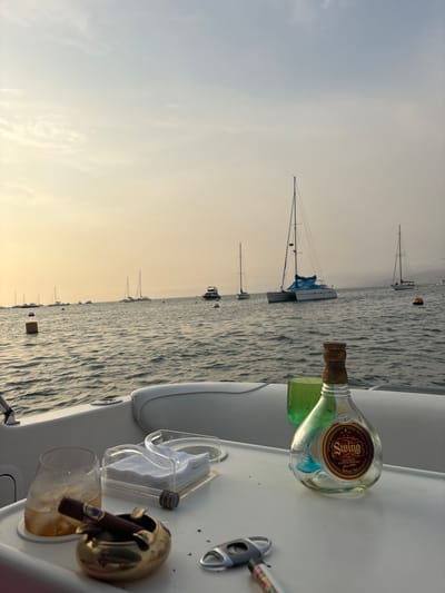 Image taken from a boat in Ancón, Peru. The scene depicts a bottle of Swing Scotch Whiskey, cigar paraphernalia, and a glass with liquor in the foreground. Sailboats are anchored in the bay under a cloudy sky.