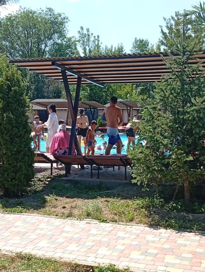 In the city of Zhibek Zholy, Kazakhstan, a recreational area features a swimming pool with several individuals. Under a wooden-slatted canopy, people are seen near the pool's edge, some standing and others seated on benches. A few individuals are in the pool itself, indicated by ripples on the water's surface. The environment includes lush green trees and bushes, and a paved walkway with a colorful brick pattern. The sky above is clear and blue.