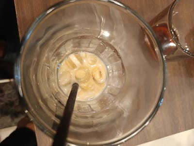 An overhead shot shows an almost empty glass of iced coffee. Cubes of ice float in a small amount of coffee liquid. A dark-colored straw sticks out. The glass is large and clear with a thick rim. Another glass, partly visible, sits next to it on a wooden table. The scene is brightly lit.